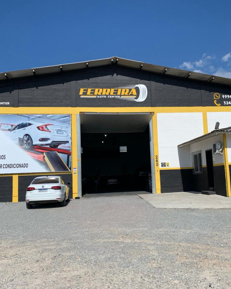 Fachada da Ferreira Auto Center, galpão grafite com faixas amarelas, a logo no alto e o portão da oficina aberto